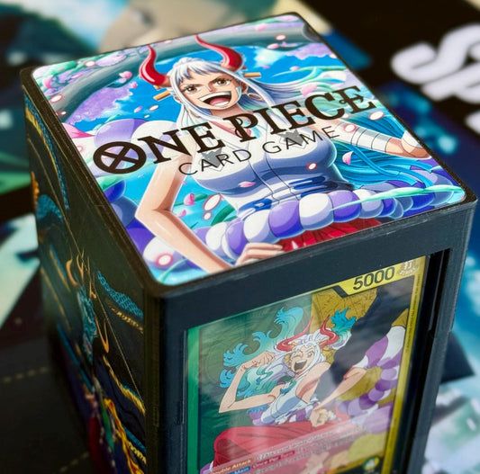 May include: A black card game deck box with a colorful image of a female anime character with long blue hair and red horns on the top. The image is from the One Piece Card Game. The box has a clear window on the front showing a card with a picture of a female anime character with long blue hair and red horns. The card has the text '5000' on it.