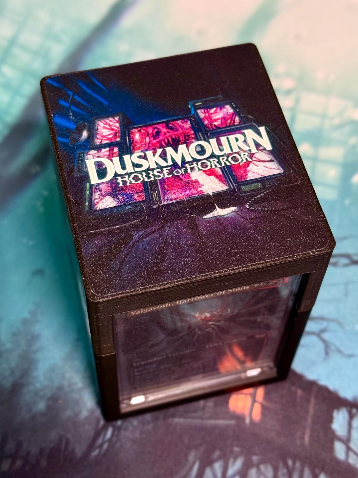 May include: Dark brown cube-shaped deck box with a textured surface. The box features a design of multiple screens displaying disturbing imagery, with the text 'DUSKMOUN HOUSE OF HORROR' prominently displayed in a stylized font.  The box also shows a partially visible Magic: The Gathering card, 'Valsavoth, Harrower of Souls', in a clear plastic window. The overall color scheme is dark, with shades of purple, pink, and blue accents.