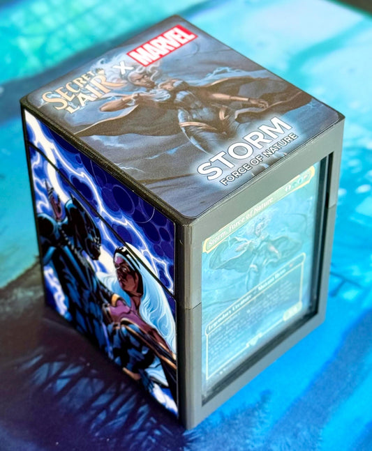 May include: A gray and black deck box with a blue and white image of Storm, a Marvel character, on the front. The box has the words 'Secret Lair X Marvel' and 'Storm Force of Nature' printed on the top. The box also has a small window on the side that shows a card with the words 'Storm Force of Nature' printed on it.