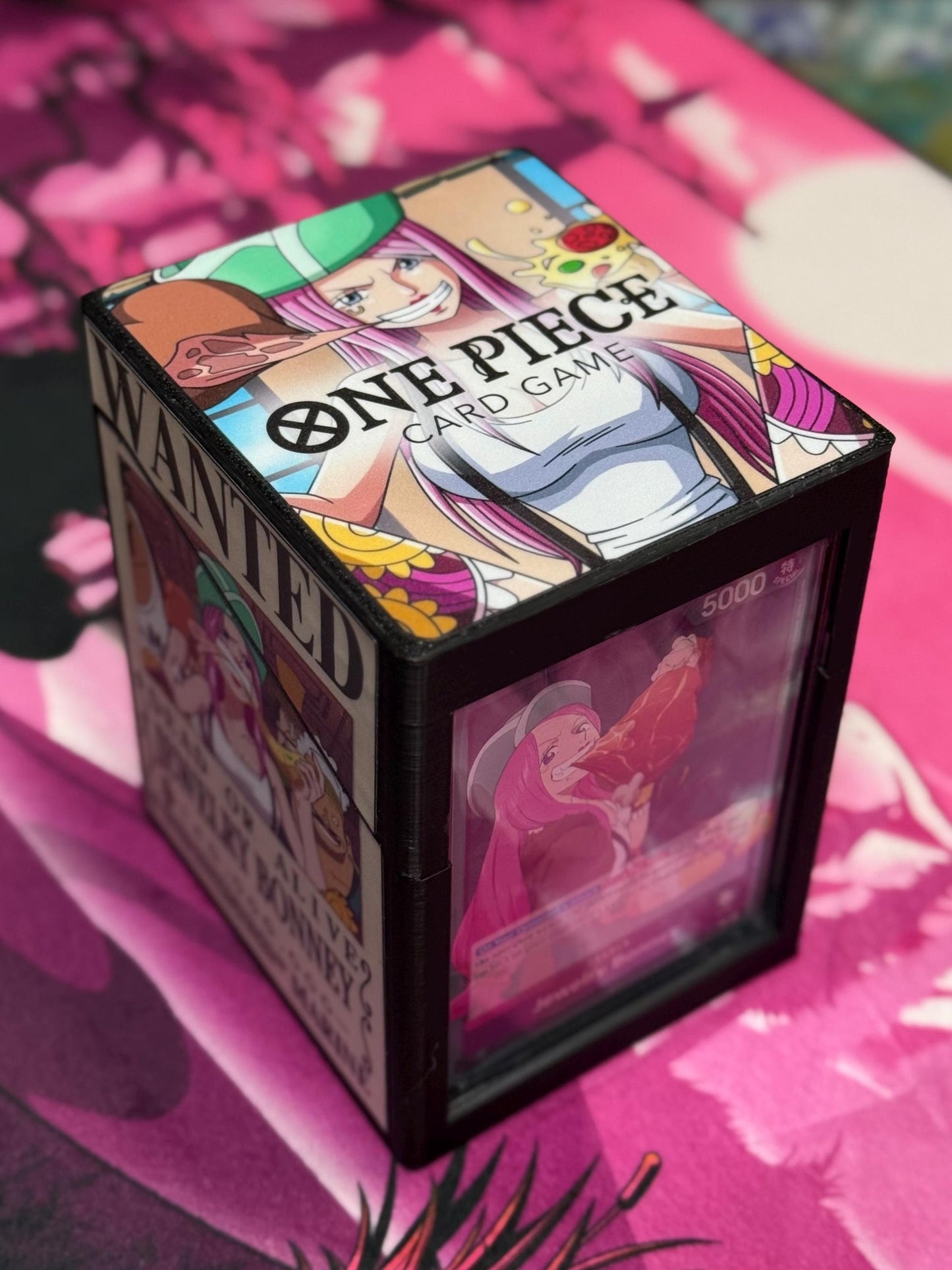 May include: A black card deck box with a clear window. The top of the box has a printed image of a pink-haired anime character with a green hat. The image says 'One Piece Card Game' and 'Wanted.' The window shows a card with a pink-haired anime character holding a chicken leg. The card has the number '5000' on it.