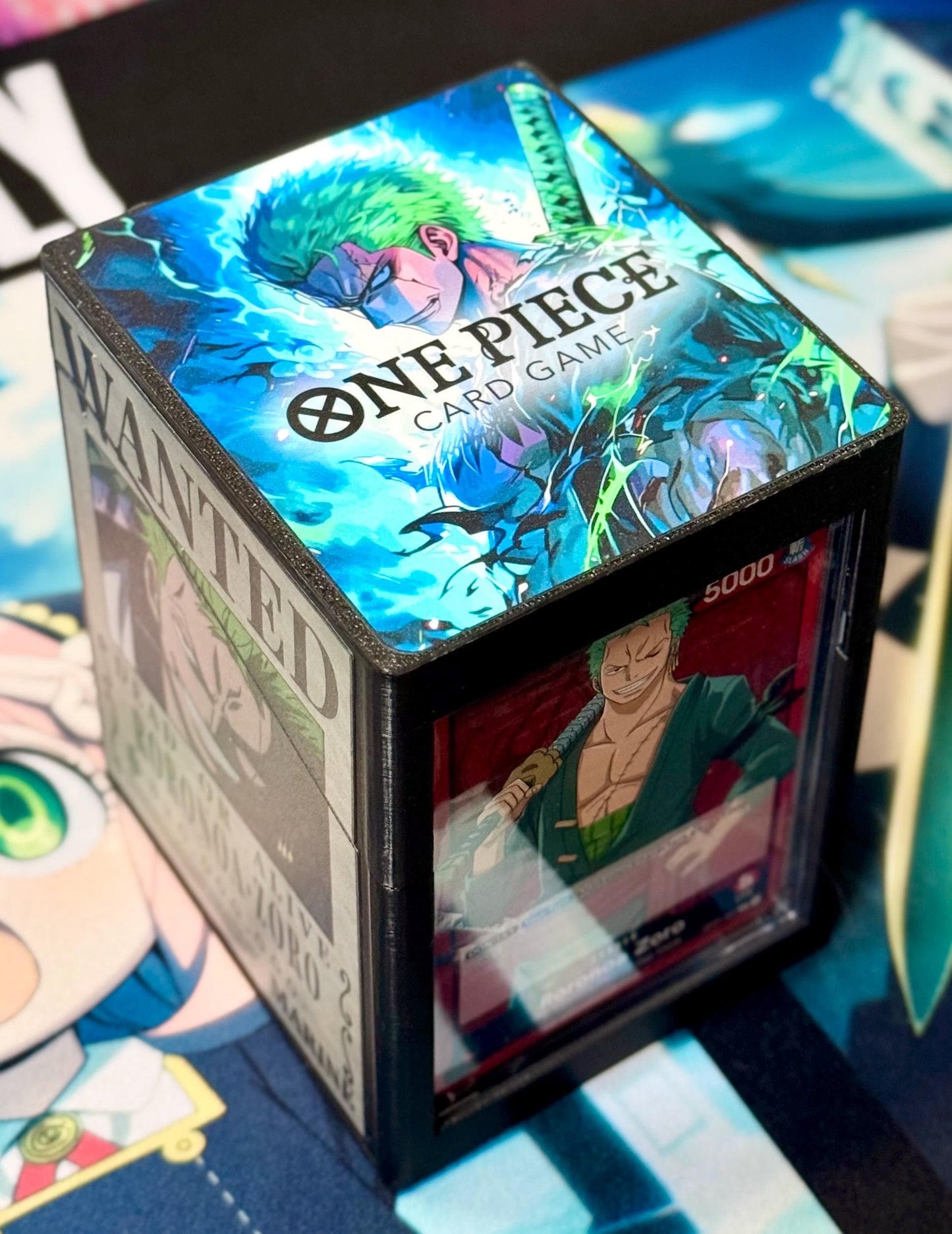May include: A black card game deck box with a clear window. The top of the box has a colorful image of a green-haired anime character with a sword. The text on the box reads 'One Piece Card Game' and 'Wanted'. The window shows a card with a red background and a green-haired anime character holding a sword. The text on the card reads '5000'.
