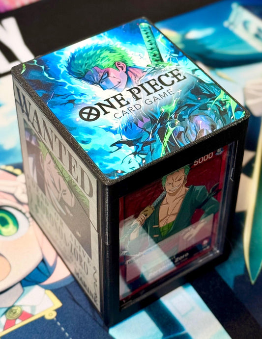 May include: A black card game deck box with a clear window. The top of the box has a colorful image of a green-haired anime character with a sword. The text on the box reads 'One Piece Card Game' and 'Wanted'. The window shows a card with a red background and a green-haired anime character holding a sword. The text on the card reads '5000'.